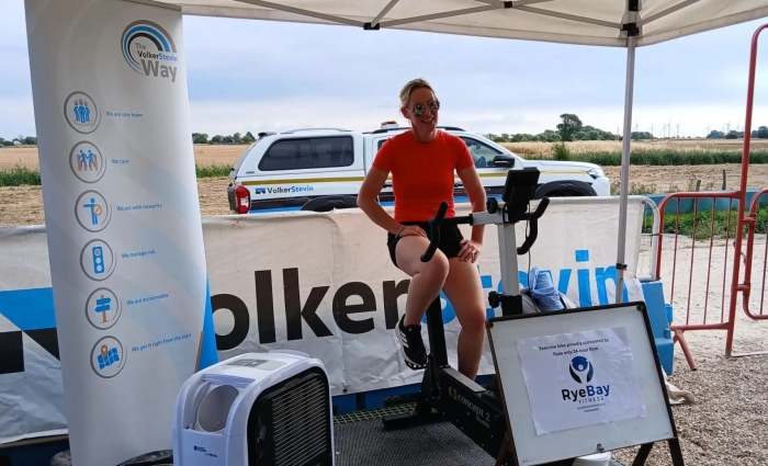 Women in black shorts and orange top on an exercise bike under a canopy