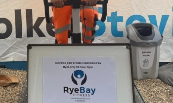 Man in orange hi vis workwear and white hard hat on an exercise bike under a canopy