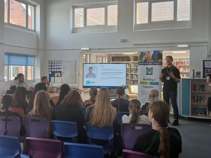 man stood in front of children in  a classroom with a presentation in a  TV screen and banner behind him