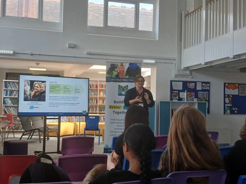 women stood in front of children in a classroom with a presentation and banner behind her